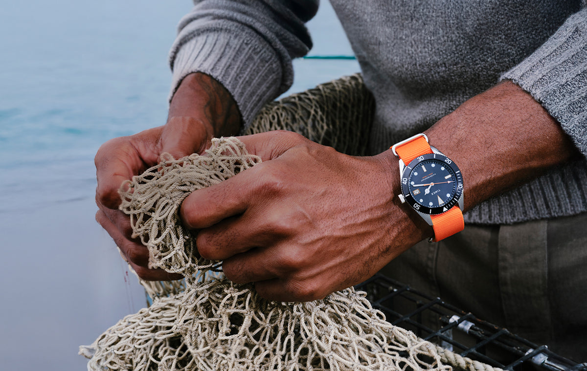 TWG069200 Man wearing a Bespoke Post x Timex Heritage 66 watch with a dark dial and bright orange fabric strap, holding a fishing net near the water.