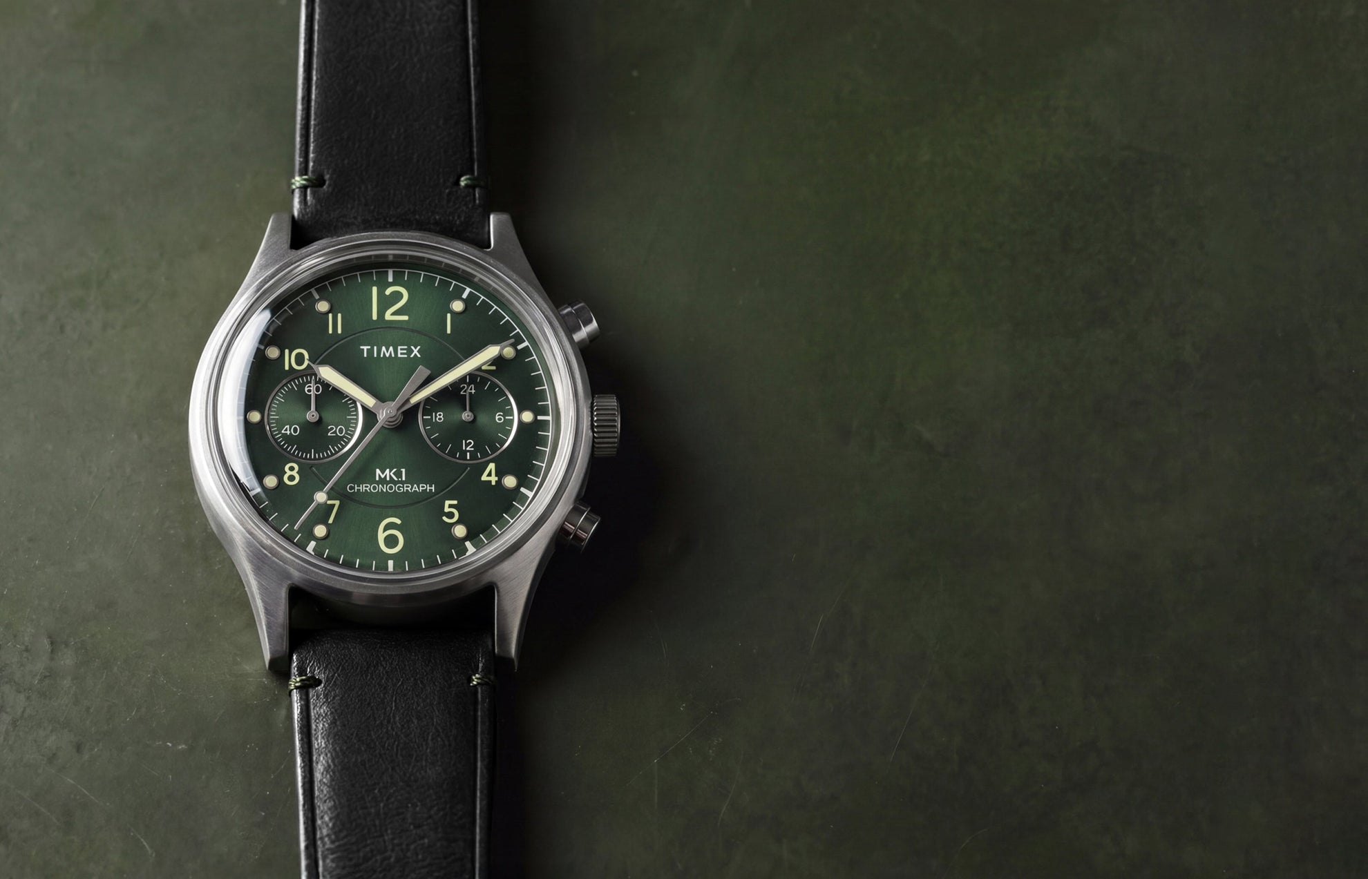 Timex MK1® Chronograph watch with a green dial, black leather strap, and brushed stainless steel case displayed on a dark green surface.