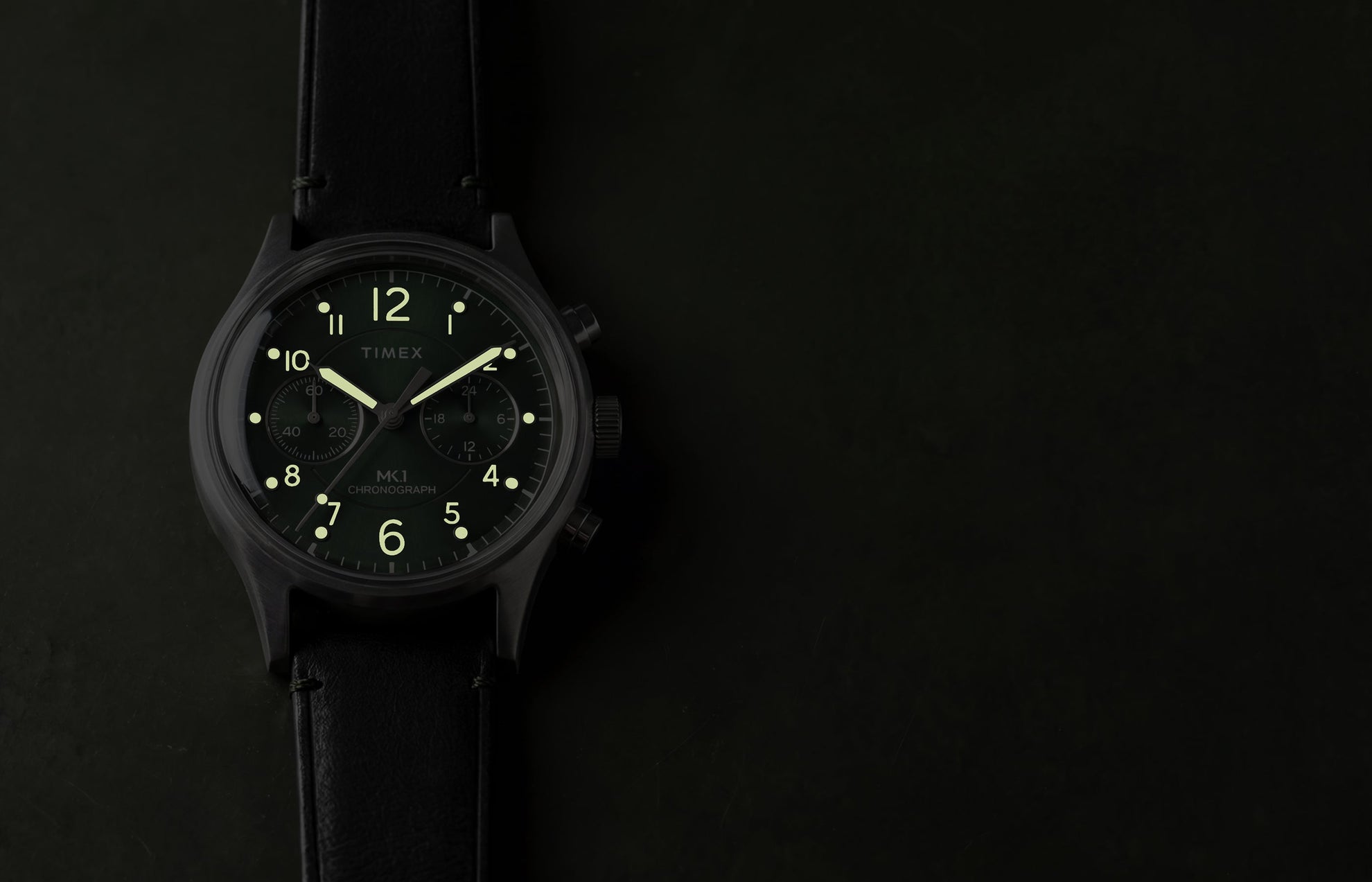 Timex MK1® Chronograph watch showing glowing luminous hands and hour markers on a green dial in low light.