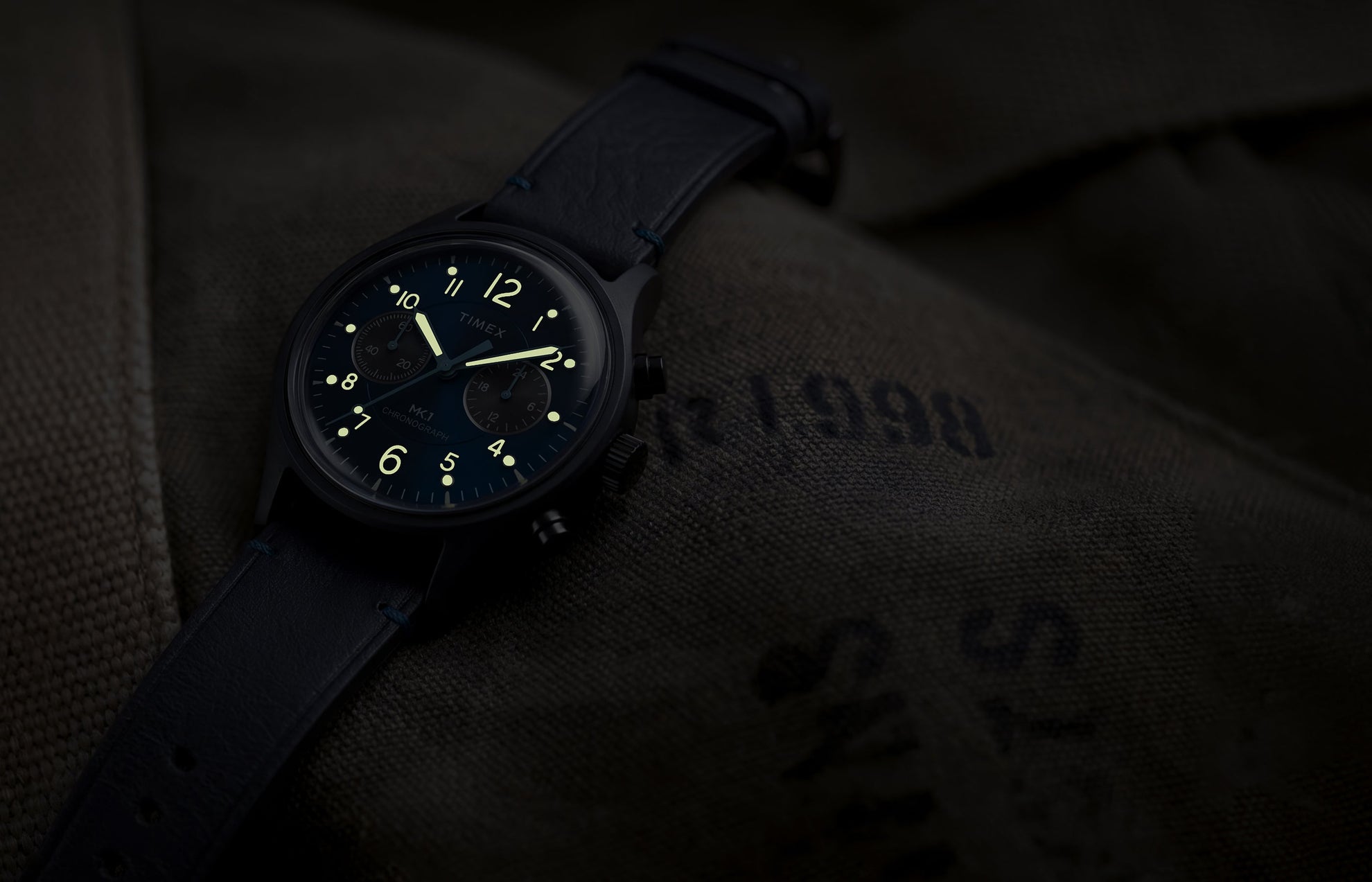 Timex MK1® Chronograph watch showing glowing luminous hands and hour markers on a blue dial in low light.