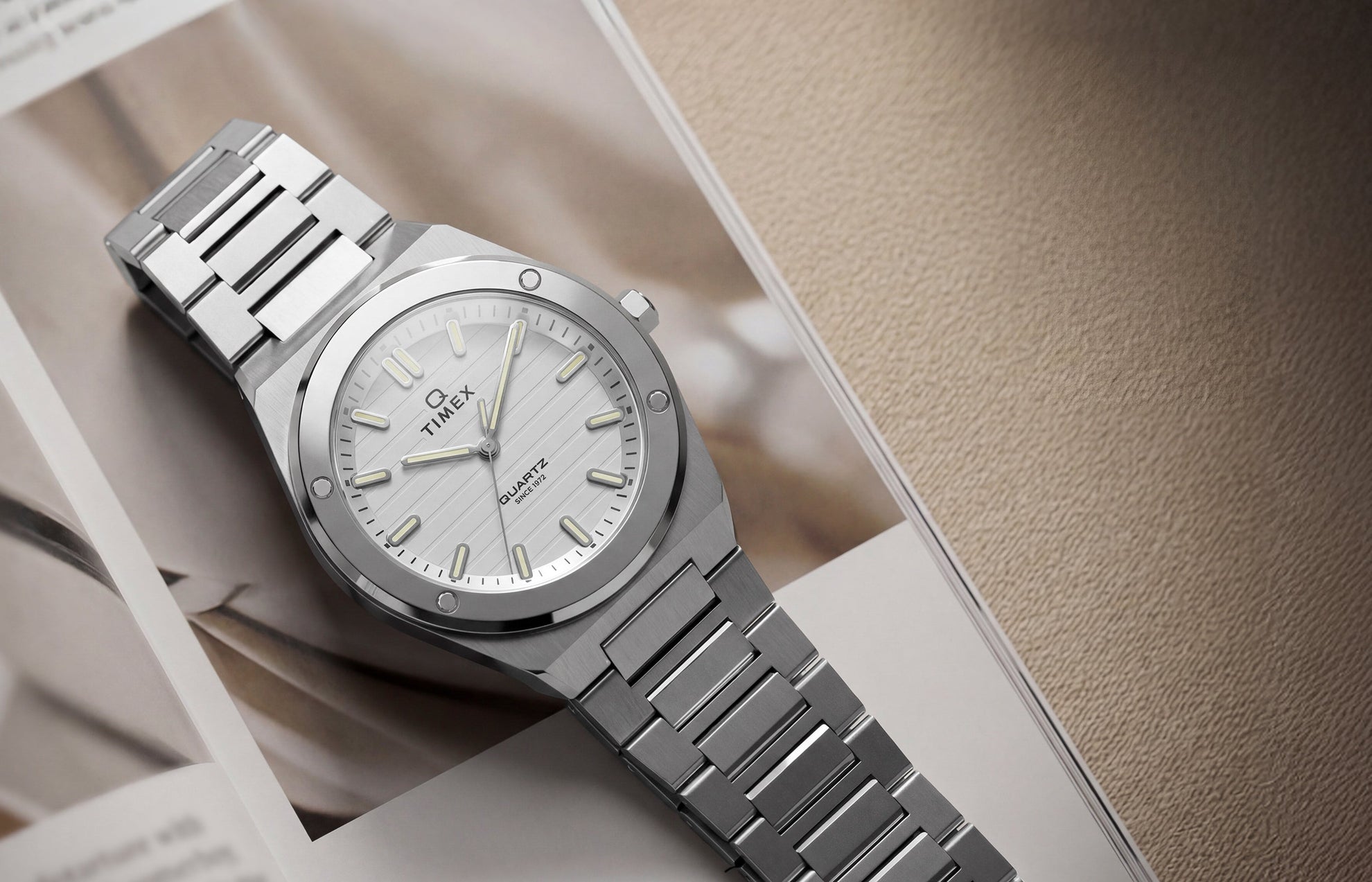 Q Timex Marbella stainless steel watch with a silver dial and integrated bracelet displayed on an open magazine.