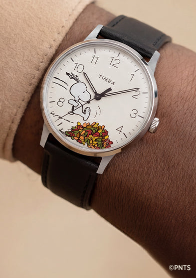 Peanuts® x Timex Waterbury Classic watch worn on the wrist, featuring a white dial, black leather strap, and Snoopy running through autumn leaves.