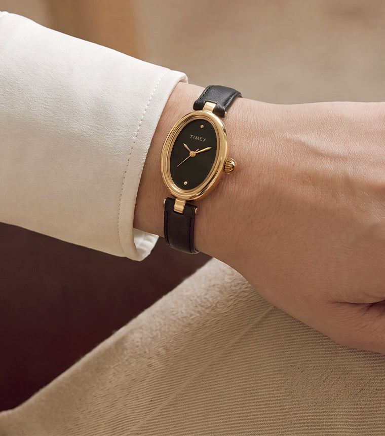 Close-up of the Timex Cavatina® Luxe with a gold-tone oval case, black dial, and black leather strap worn on the wrist.