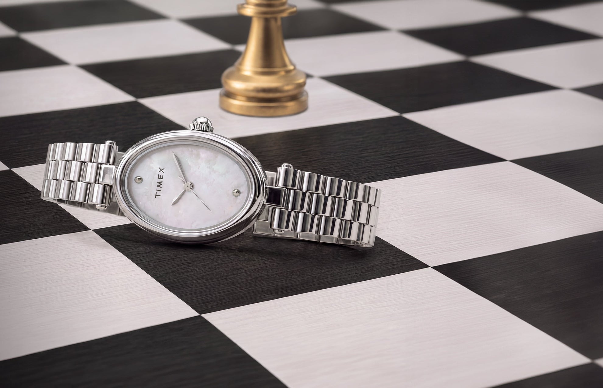 Timex Cavatina® Luxe watch with a silver-tone stainless steel bracelet, oval case, and mother-of-pearl dial displayed on a black-and-white chessboard beside a gold chess piece.