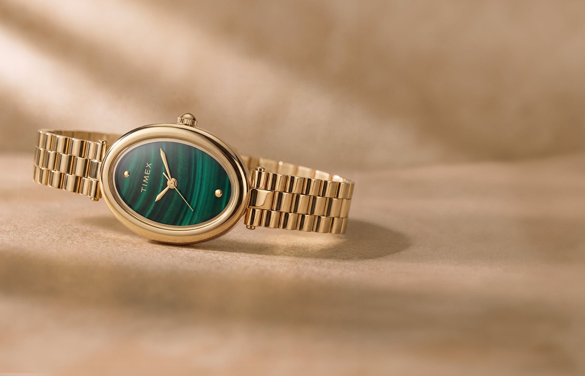 Timex Cavatina® Luxe watch with a gold-tone stainless steel bracelet, oval case, and green malachite-pattern dial displayed on a warm neutral surface.