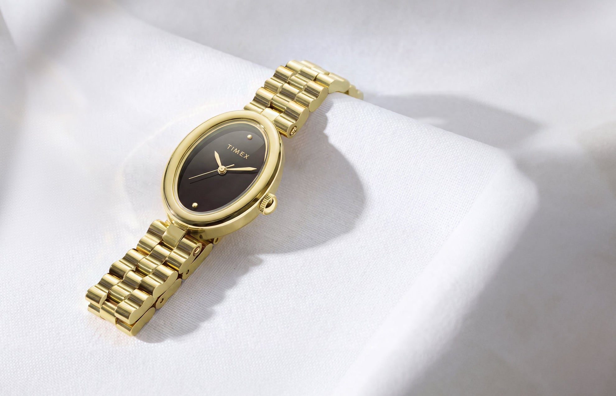 Timex Cavatina® Luxe watch with a gold-tone stainless steel bracelet, oval case, and black dial displayed on soft white fabric.