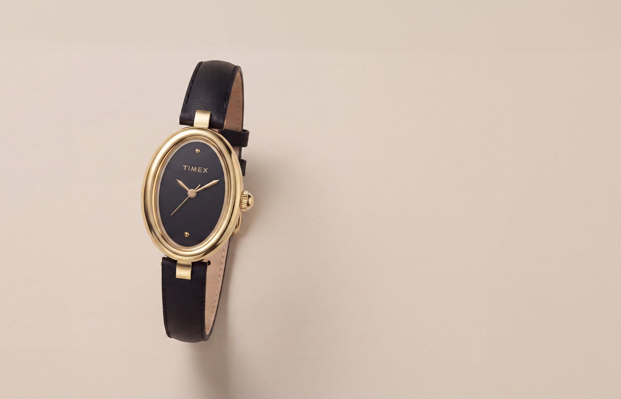 Timex Cavatina® Luxe watch with a gold-tone oval case, black dial, and black leather strap displayed on a warm neutral background.