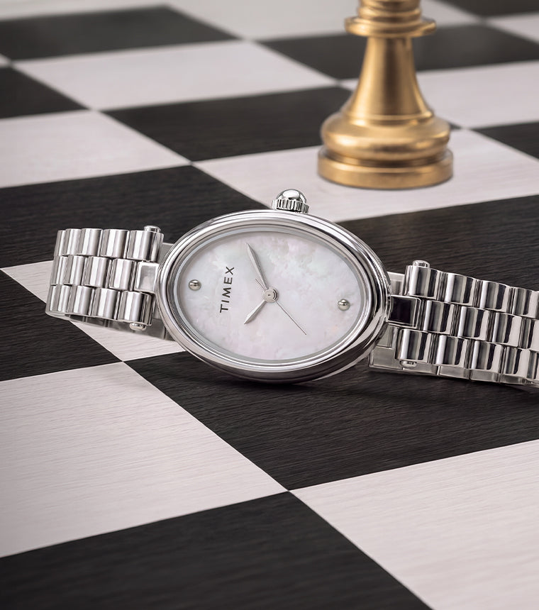 Timex Cavatina® Luxe watch with a silver-tone stainless steel bracelet, oval case, and mother-of-pearl dial displayed on a black-and-white chessboard beside a gold chess piece.
