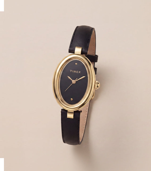 Timex Cavatina® Luxe watch with a gold-tone oval case, black dial, and black leather strap displayed on a warm neutral background.
