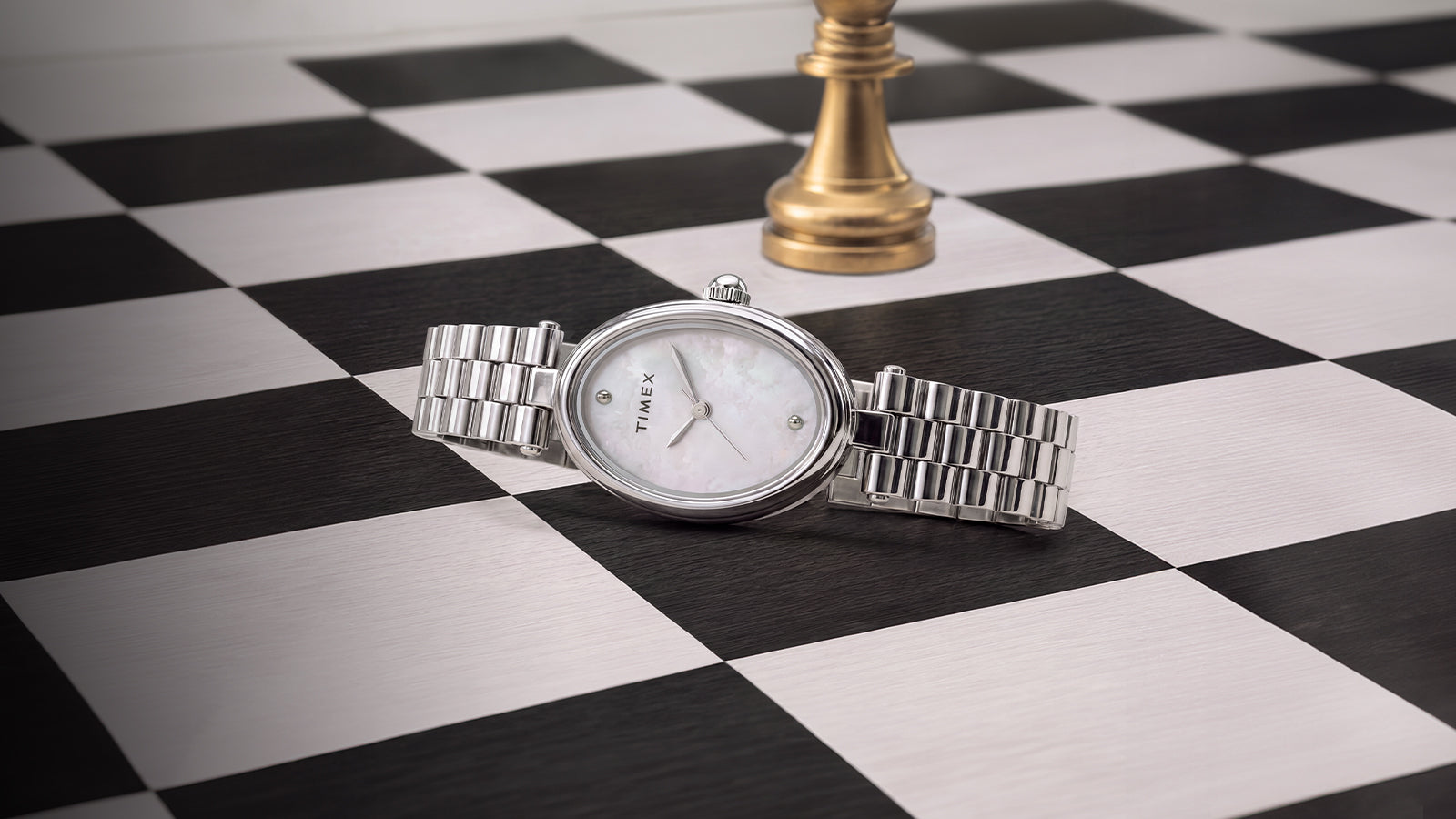 Timex Cavatina® Luxe silver-tone stainless steel watch with a mother-of-pearl dial displayed on a black-and-white chessboard with a gold chess piece in the background.