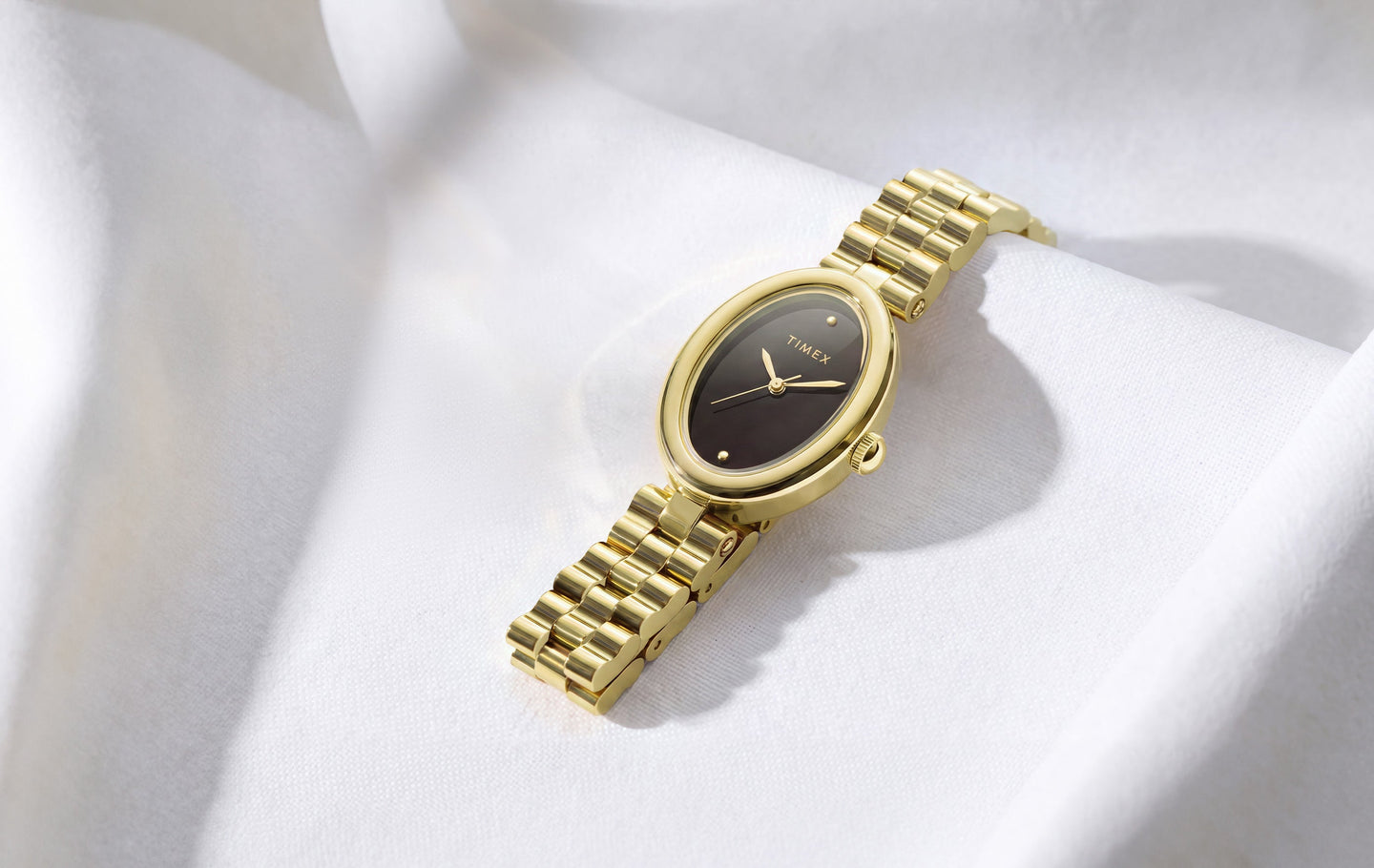 Gold-tone Timex Cavatina® Luxe watch with a dark oval dial and polished metal bracelet, displayed on softly draped white fabric.
