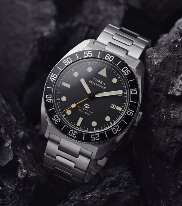 Timex Expedition Pioneer Automatic with a black dial, black rotating bezel, and stainless-steel bracelet displayed among dark charcoal rocks.