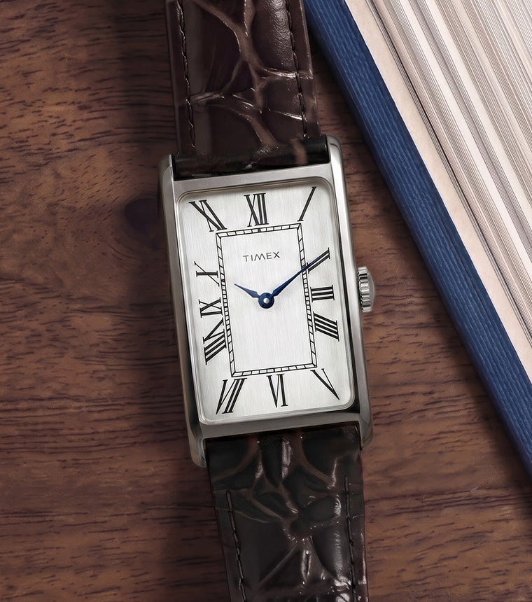 Timex 1976 Lexington Reissue Inspired rectangular watch with a brushed silver-tone dial, black Roman numeral hour markers, blue hands, and a brown crocodile-pattern leather strap, resting on a wooden tabletop beside a blue hardcover book.