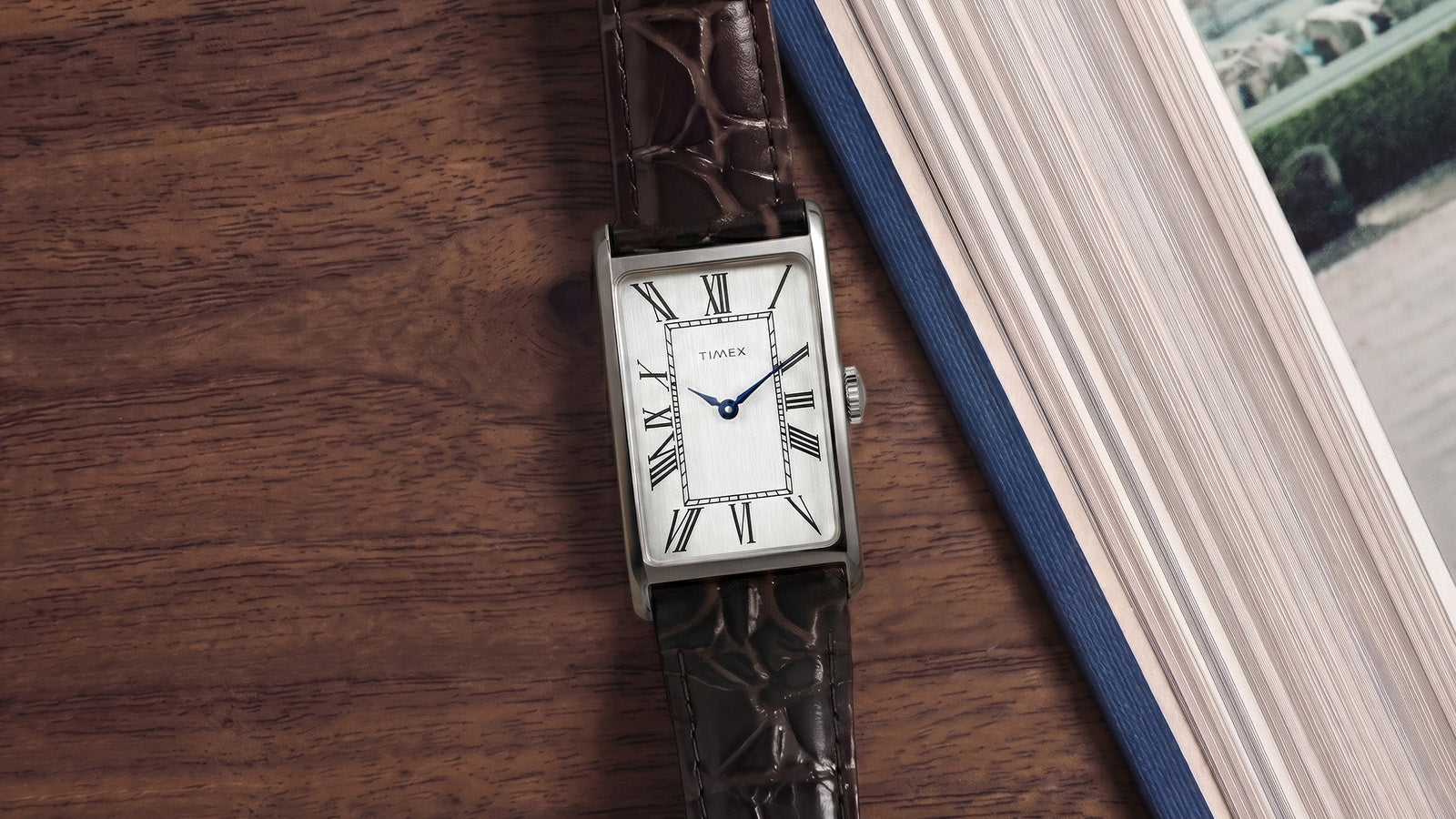Timex 1976 Lexington Reissue Inspired watch displayed beside an open book, showcasing its rectangular silver-tone case, Roman numeral dial, and brown leather strap.