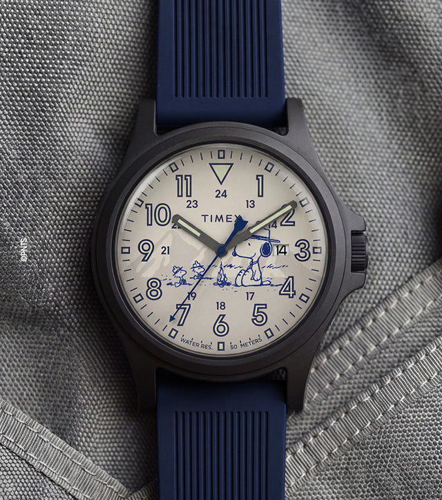 Peanuts® x Timex Expedition® Acadia watch with a matte black case, cream dial, and navy blue silicone strap, featuring Snoopy with Woodstock artwork, bold blue Arabic numerals, a date window, and blue hands displayed on a gray tactical fabric background.