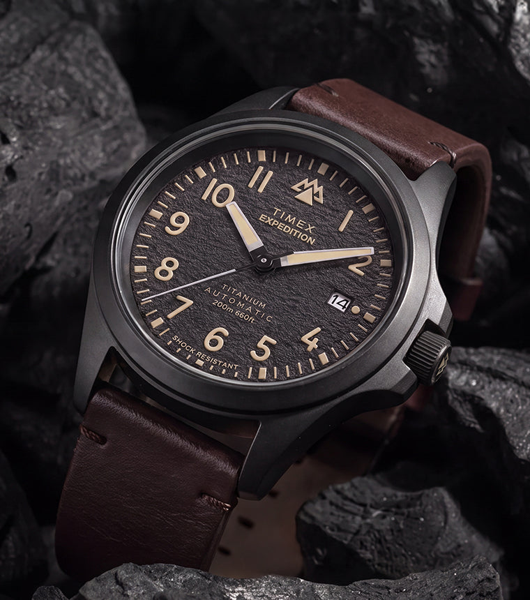 Expedition® Field Post Automatic titanium watch with a black dial and brown leather strap displayed among charcoal rocks.