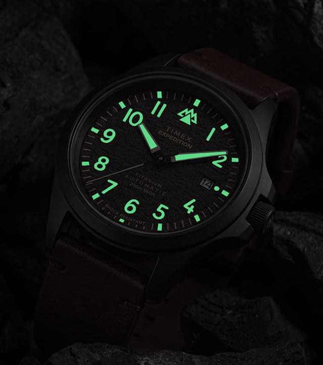 Expedition® Field Post Automatic titanium watch with glowing luminous hands and hour markers in low light.