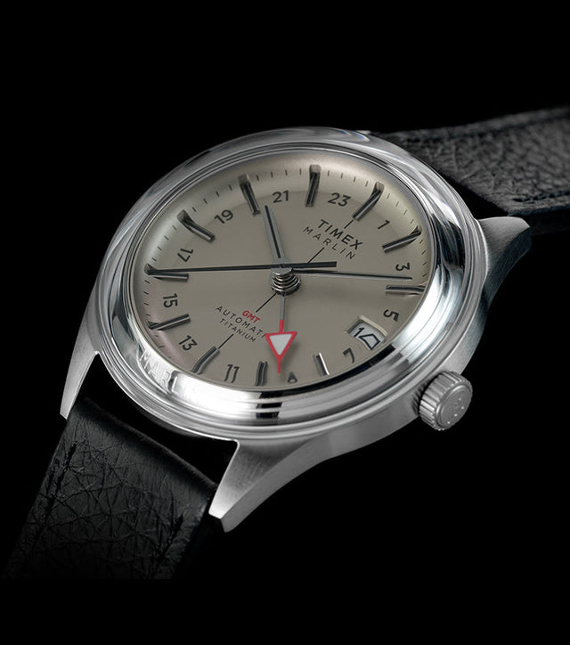 Timex Marlin® Jet Titanium Automatic GMT watch with silver-tone titanium case, black leather strap, cream dial, red GMT hand, and date window on a black background.
