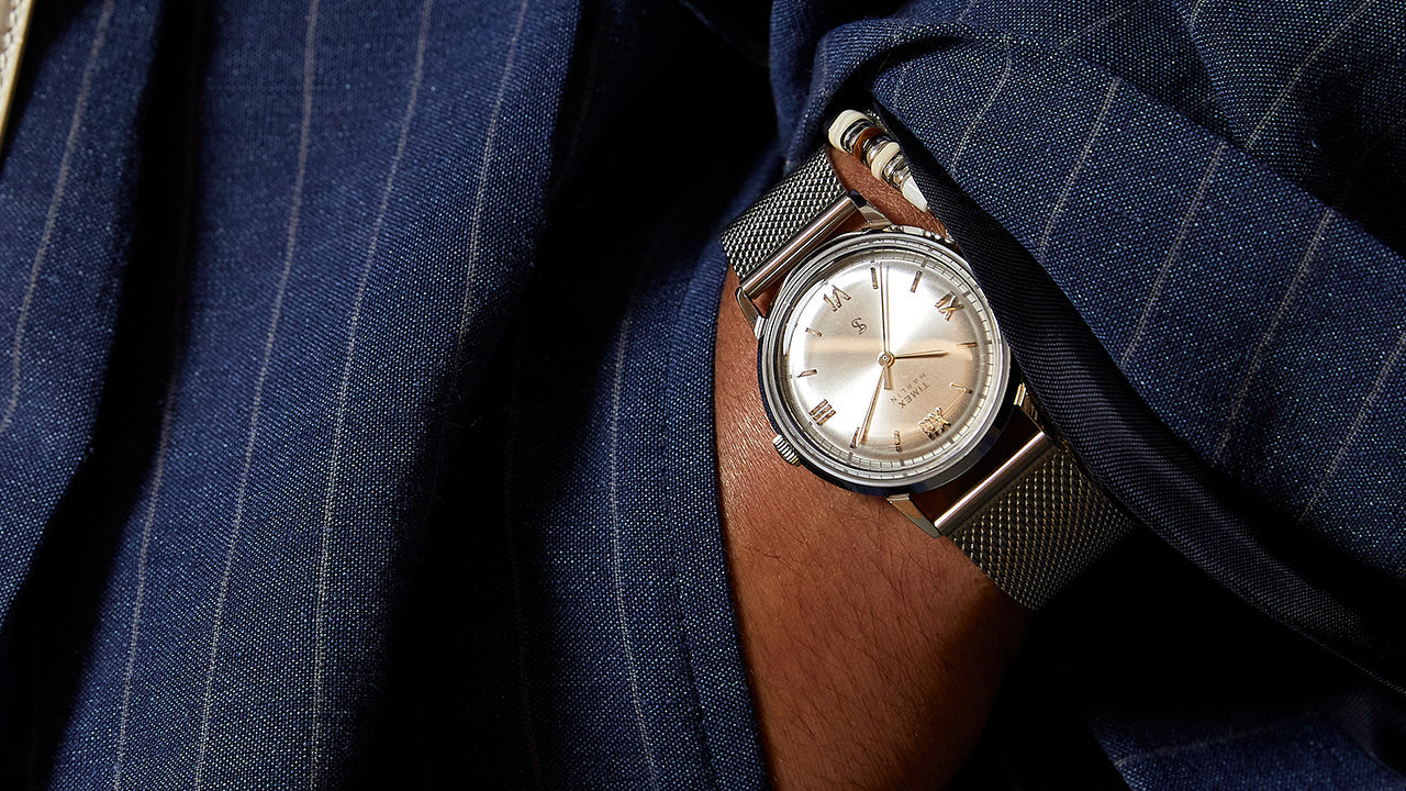 Todd Snyder x Timex Marlin® Mesh watch with a silver sunray dial worn beneath the cuff of a navy pinstripe suit jacket.