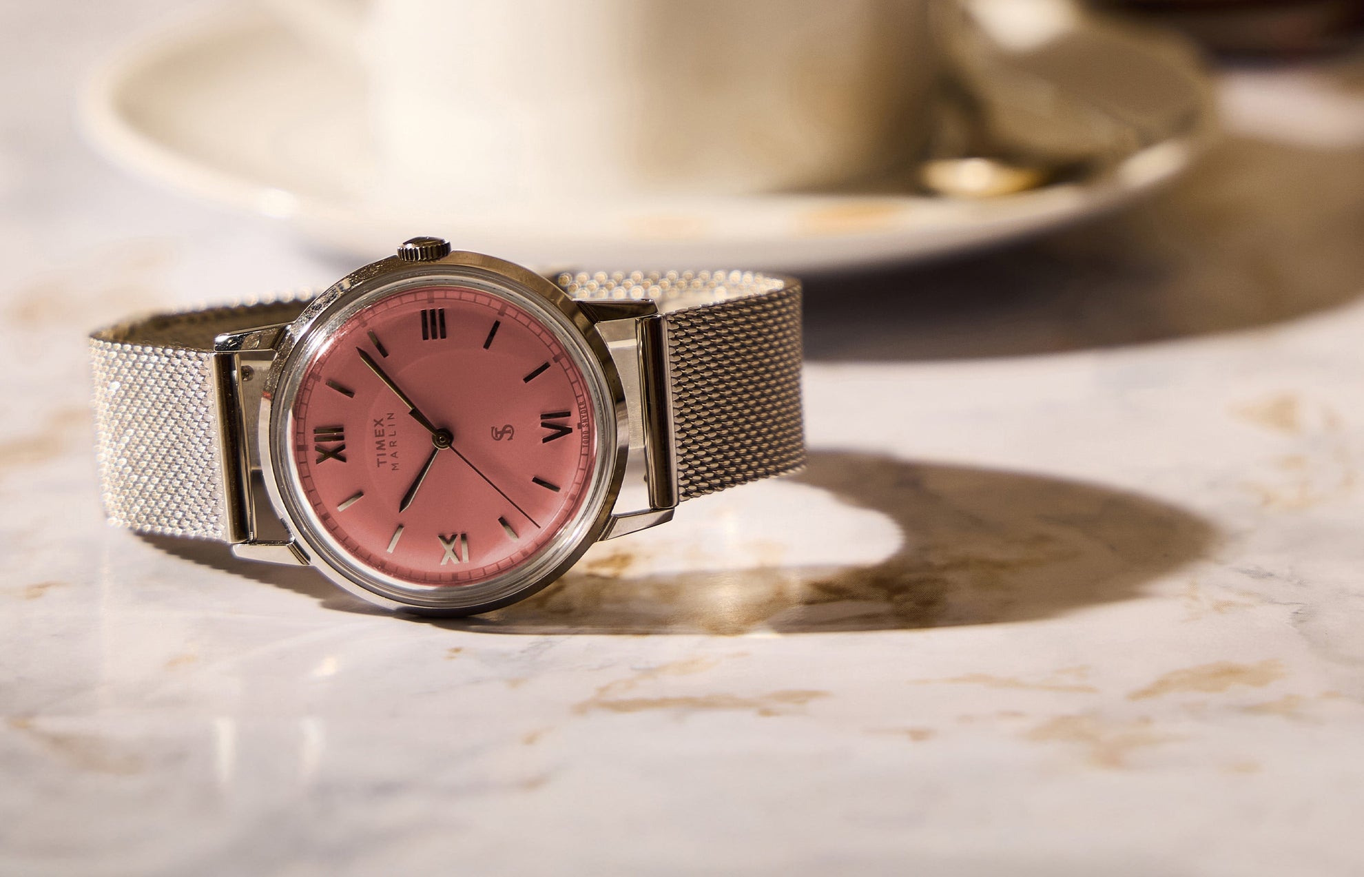 Todd Snyder x Timex Marlin® Mesh watch with a pink dial and stainless steel mesh bracelet displayed on a marble table beside a coffee cup.