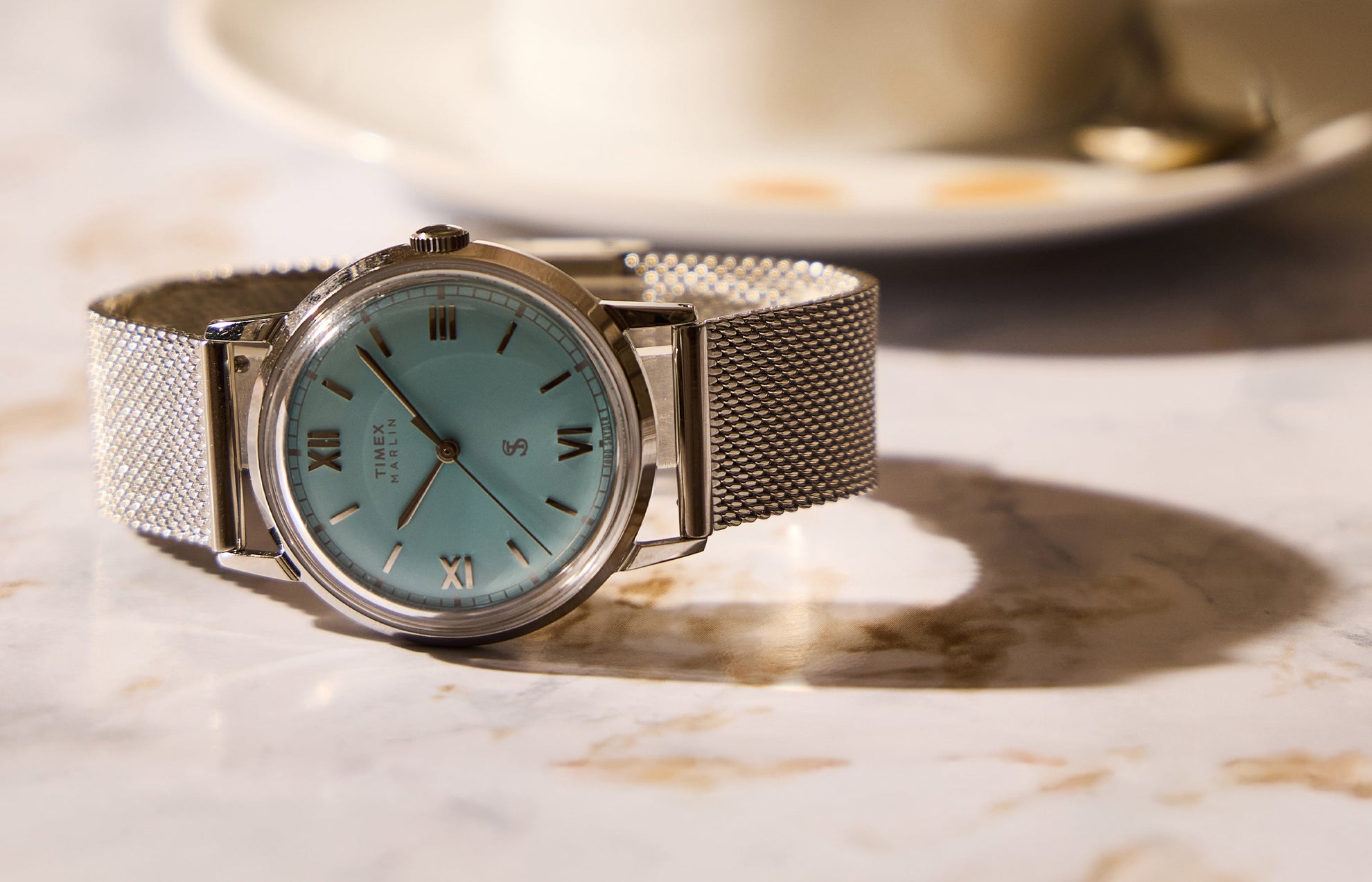 Todd Snyder x Timex Marlin® Mesh watch with a light blue dial and stainless steel mesh bracelet displayed on a marble table beside a coffee cup.
