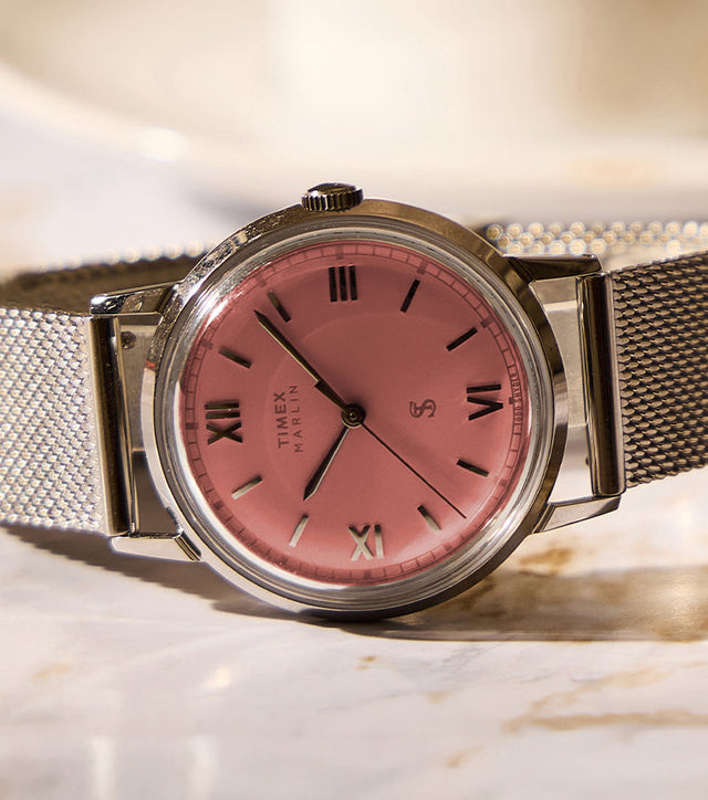 Todd Snyder x Timex Marlin® Mesh watch with a pink dial and stainless steel mesh bracelet displayed on a marble table beside a coffee cup.