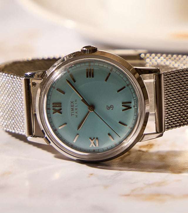 Todd Snyder x Timex Marlin® Mesh watch with a light blue dial and stainless steel mesh bracelet displayed on a marble table beside a coffee cup.