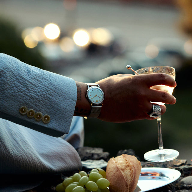 Person wearing the Todd Snyder x Timex Marlin® Mesh watch while holding a cocktail glass during an outdoor dining setting.