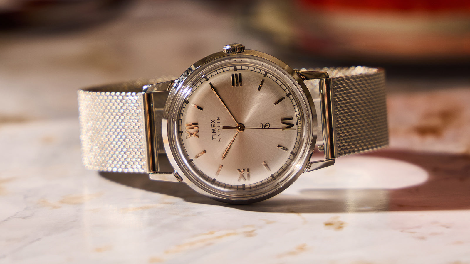Todd Snyder x Timex Marlin® Mesh watch with a silver sunray dial, Roman numeral hour markers, and a stainless steel mesh bracelet on a marble surface.
