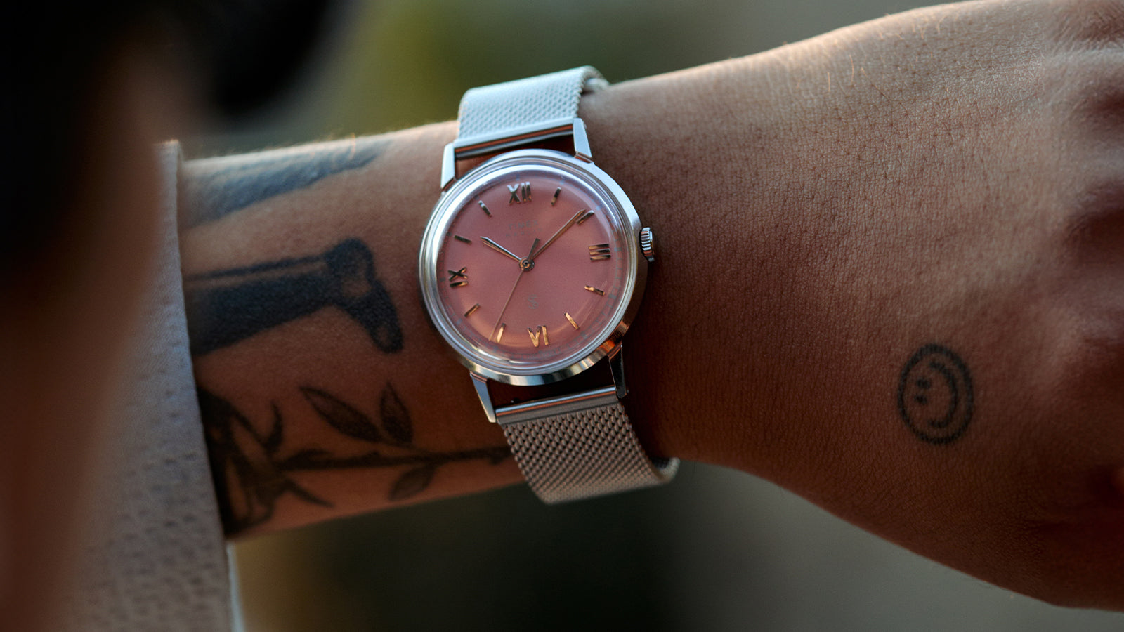 Detailed view of the Todd Snyder x Timex Marlin® Mesh watch featuring a pink dial, Roman numeral hour markers, and a stainless steel mesh bracelet on the wrist.