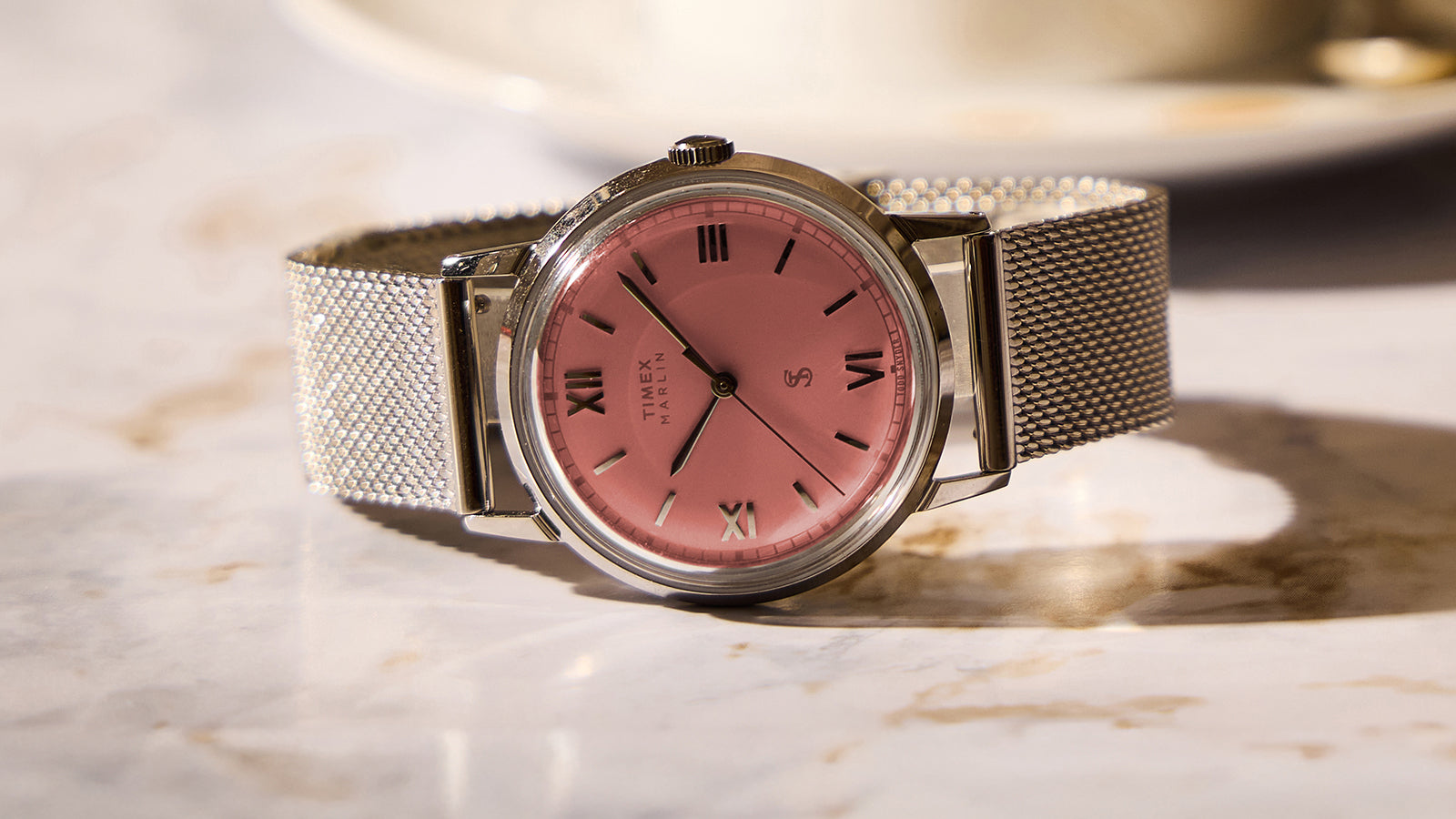 Todd Snyder x Timex Marlin® Mesh watch with a pink dial, Roman numeral hour markers, and a stainless steel mesh bracelet on a marble surface.