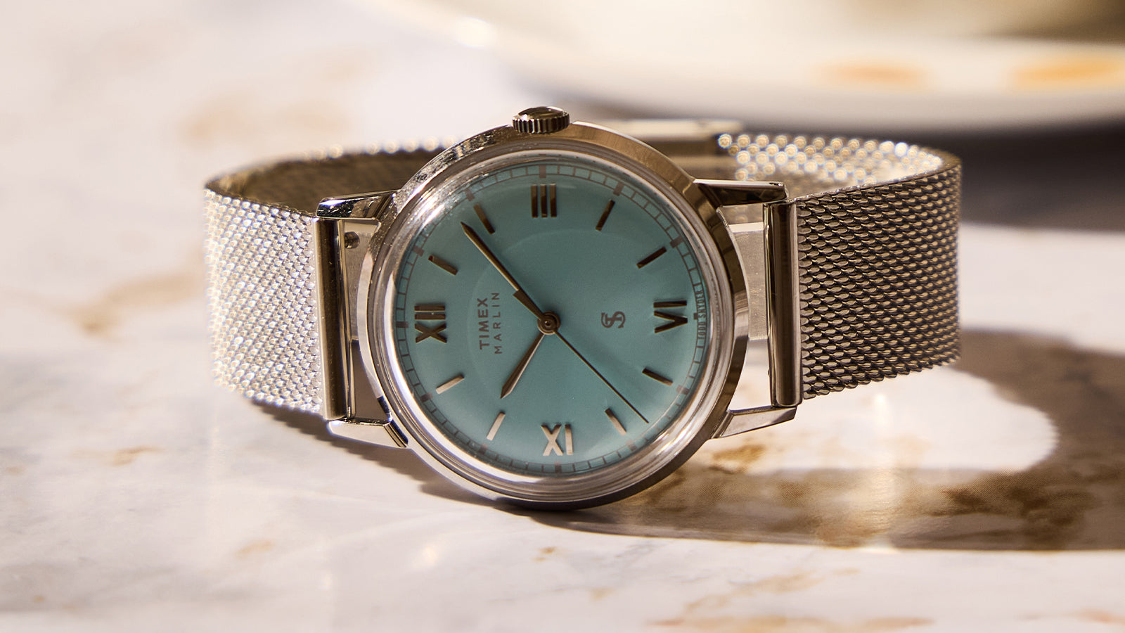 Todd Snyder x Timex Marlin® Mesh watch with a light blue dial, Roman numeral hour markers, and a stainless steel mesh bracelet on a marble surface.