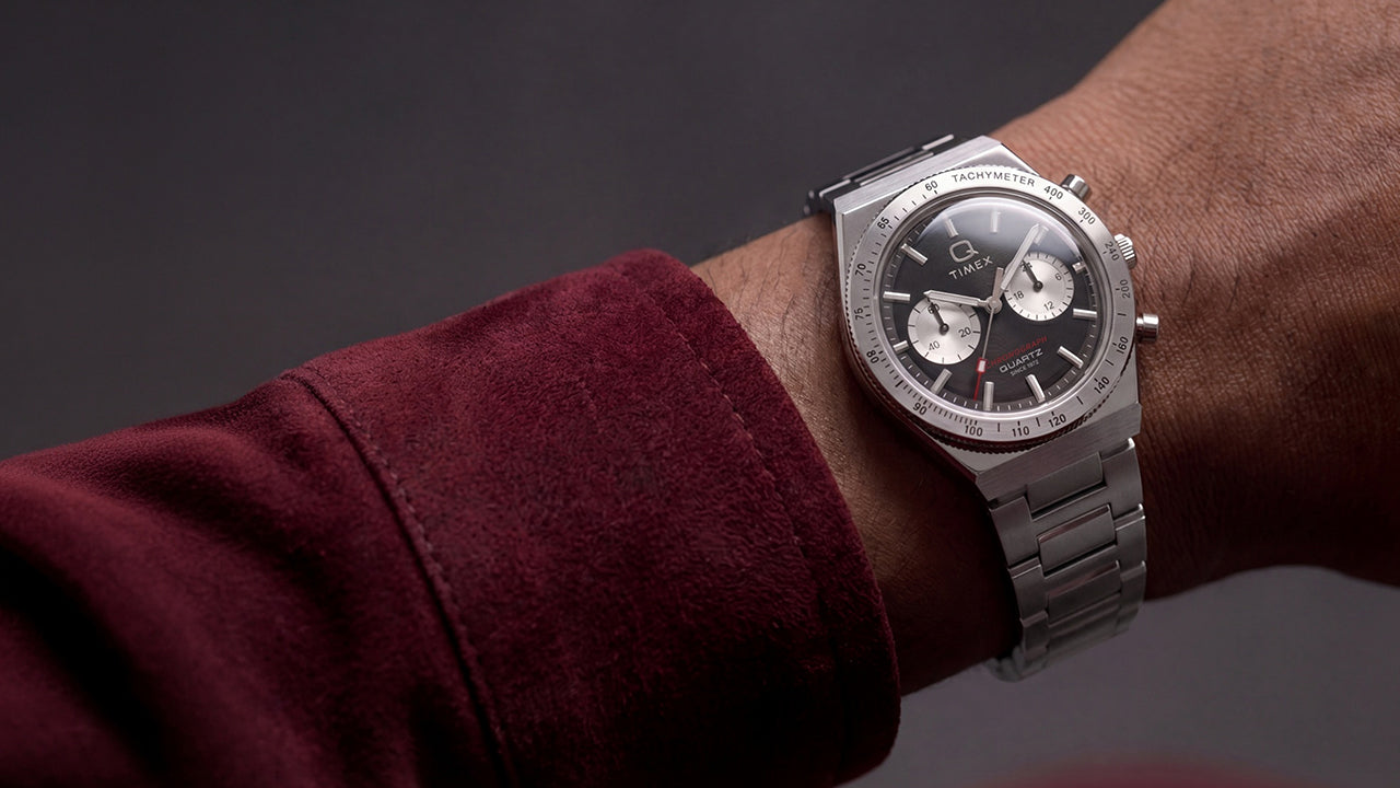 Q Timex Continental Chronograph worn on the wrist with a burgundy jacket, featuring a black chronograph dial and stainless-steel bracelet.