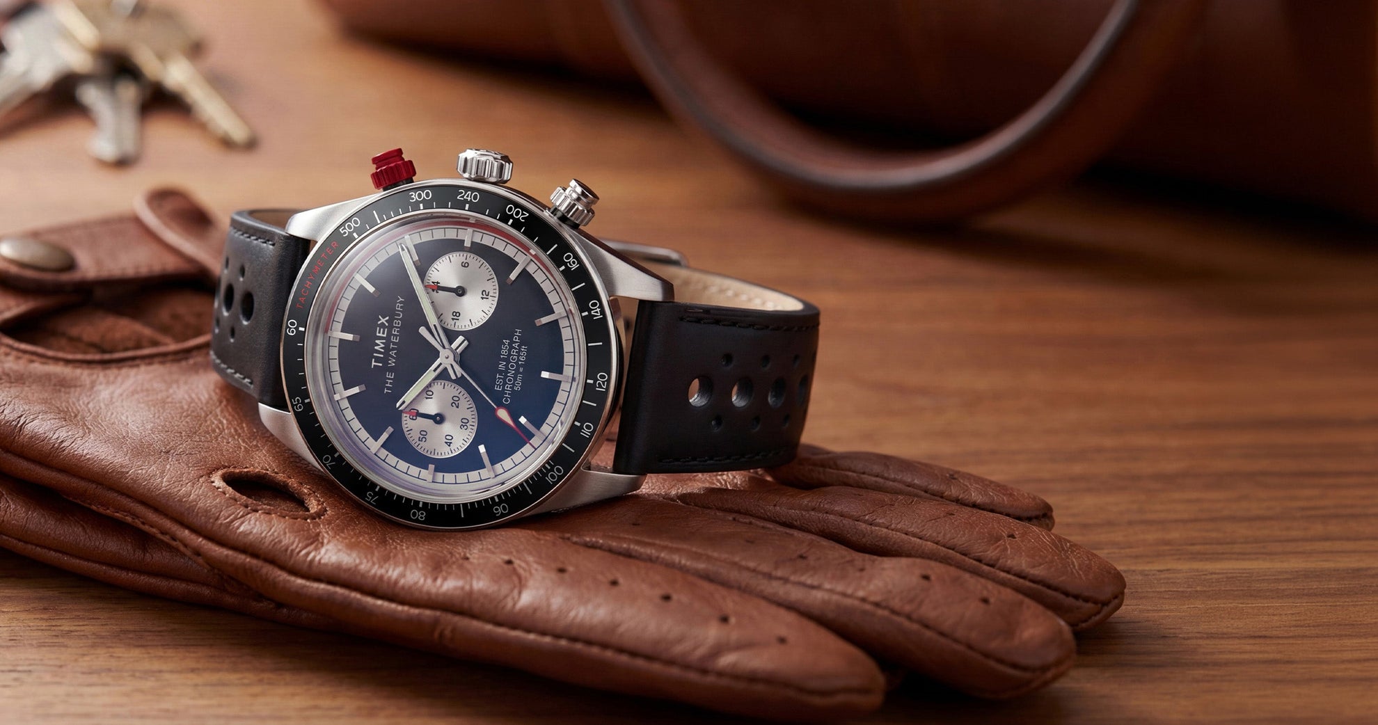 Timex Waterbury Heritage Chronograph watch with a black leather racing strap, black tachymeter bezel, panda-style chronograph dial, and red chronograph pusher displayed on brown driving gloves.
