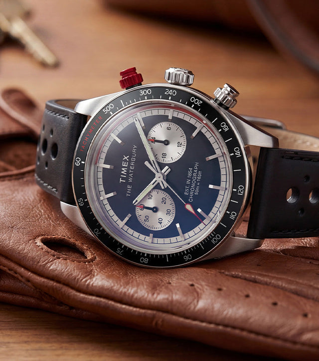 Timex Waterbury Heritage Chronograph watch with a black leather racing strap, black tachymeter bezel, panda-style chronograph dial, and red chronograph pusher displayed on brown driving gloves.