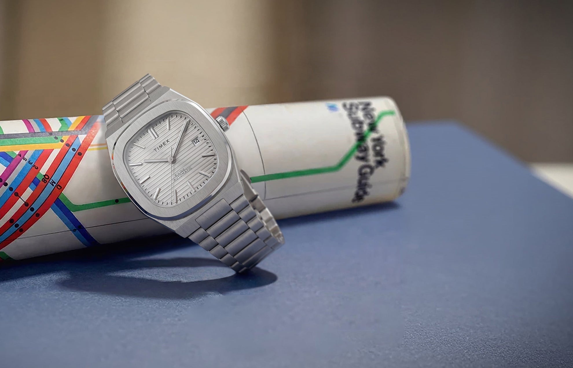 Timex® Automatic E Line 38mm watch with a stainless steel bracelet, silver textured dial, and date window displayed on a rolled New York City subway map.