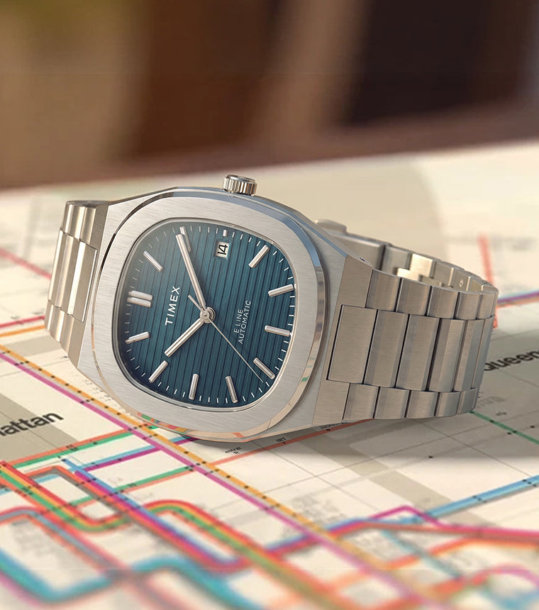 Timex® Automatic E Line 38mm watch with a stainless steel bracelet, blue textured dial, and date window displayed on a transit map.