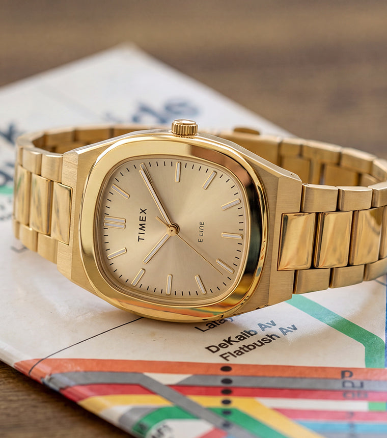 Timex® Quartz E Line 30mm watch with a gold-tone stainless steel bracelet and champagne dial displayed on a New York City subway map.