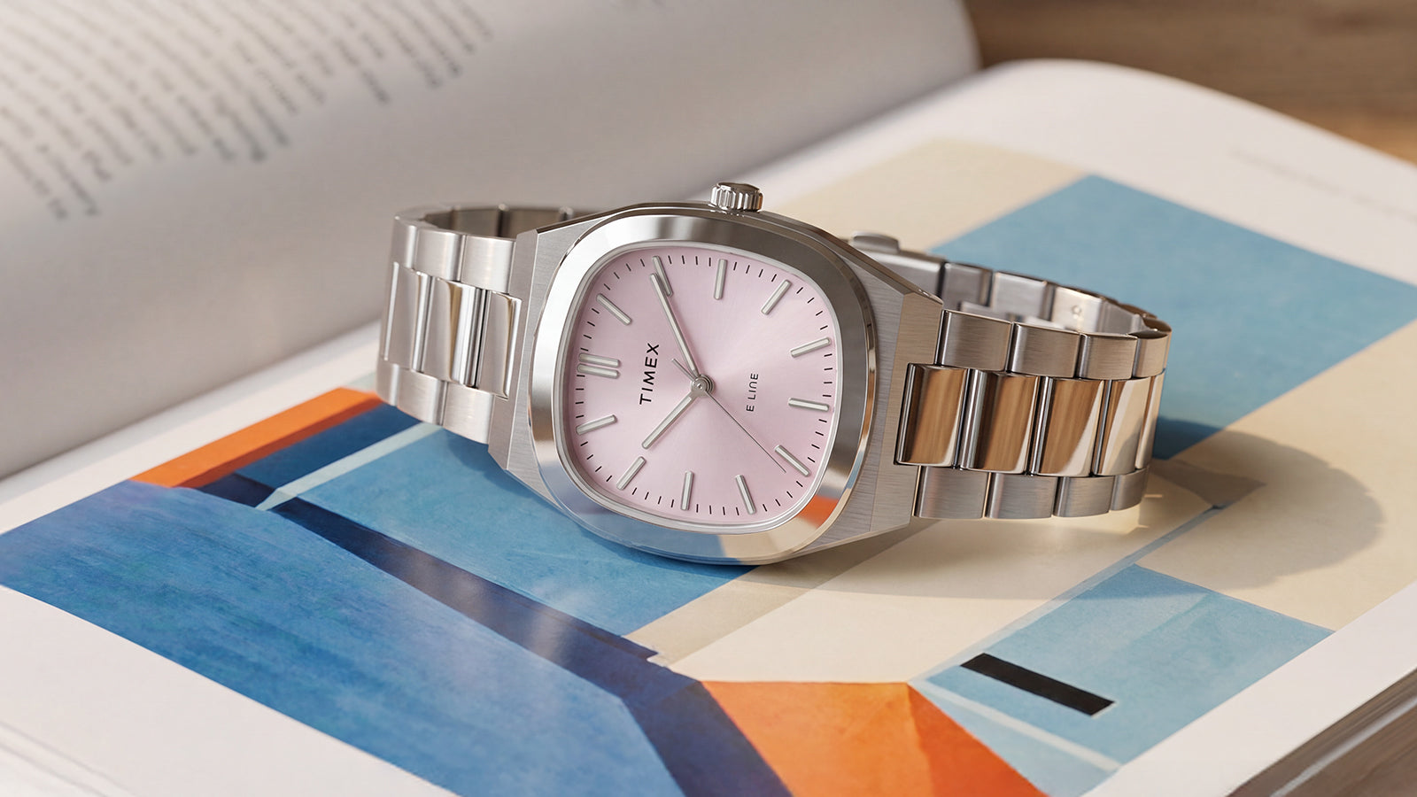 Close-up of the Timex® Quartz E Line 30mm stainless steel watch with a pink dial displayed on an open book with colorful artwork.