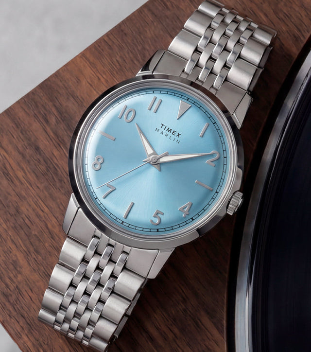Timex Marlin Quartz watch with an ice blue sunray dial and stainless steel bracelet displayed on a wooden tabletop beside a vinyl record.