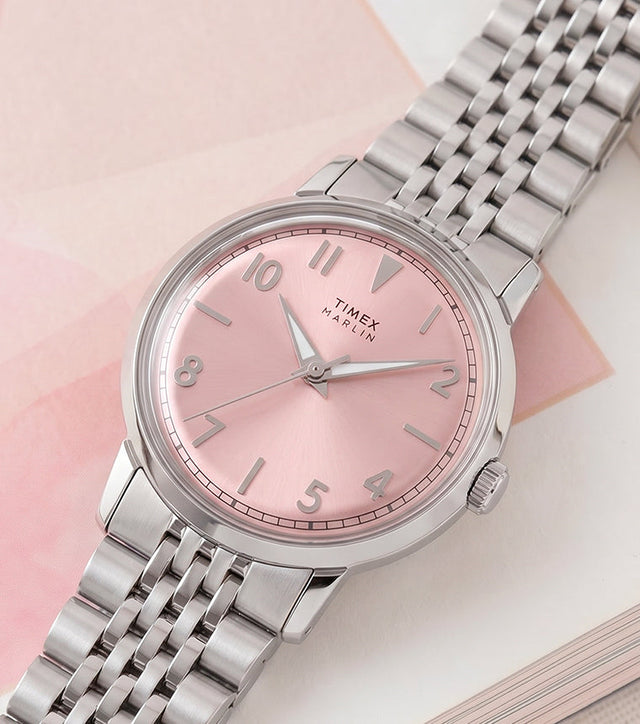 Timex Marlin Quartz watch with a pink sunray dial and stainless steel bracelet displayed on an open book with soft pink accents.