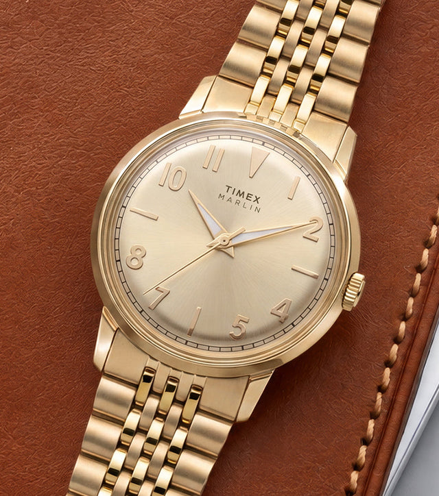 Timex Marlin Quartz watch with a champagne sunray dial and gold-tone stainless steel bracelet displayed on a leather portfolio.
