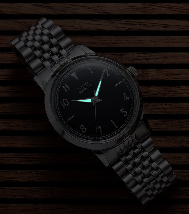 Close-up of the Timex Marlin Quartz with luminous hands illuminated on a black sunray dial in low light.