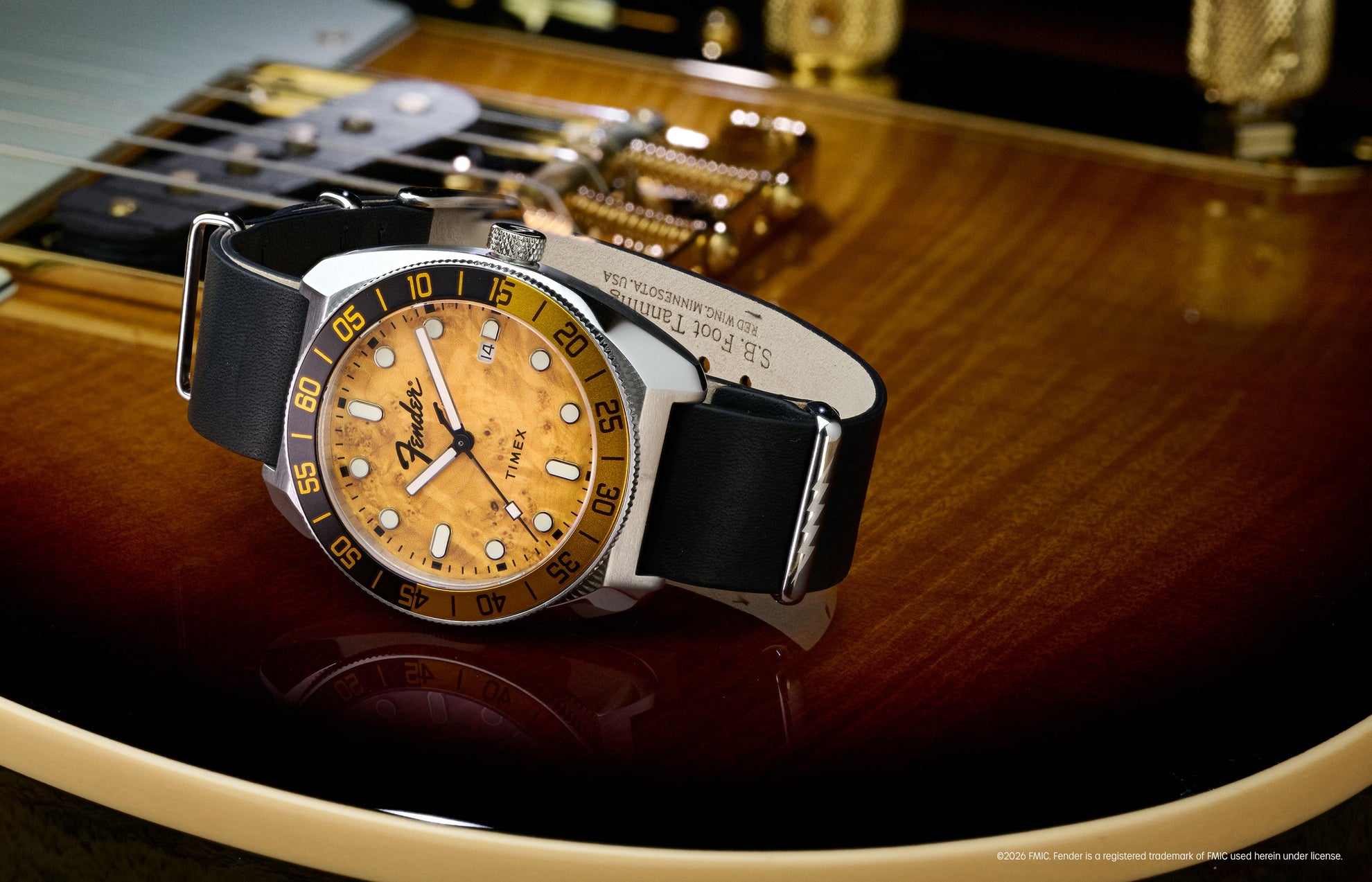 Fender x Timex Automatic watch with a silver-tone case, amber wood-grain dial, black-and-gold bezel, and black leather strap displayed on a Fender guitar.