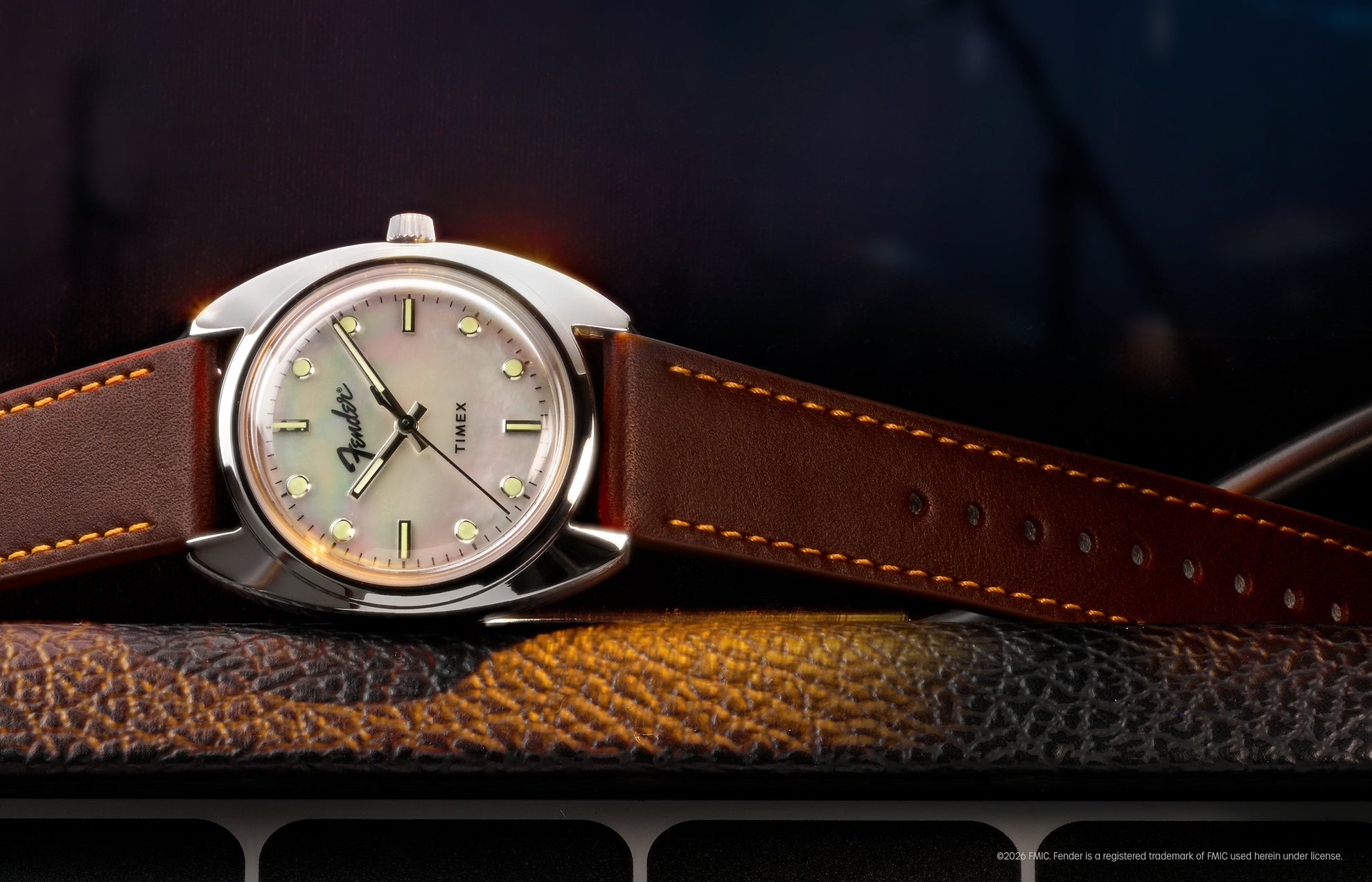 Fender x Timex watch with a silver-tone case, iridescent mother-of-pearl dial, and brown leather strap displayed on a guitar amplifier.