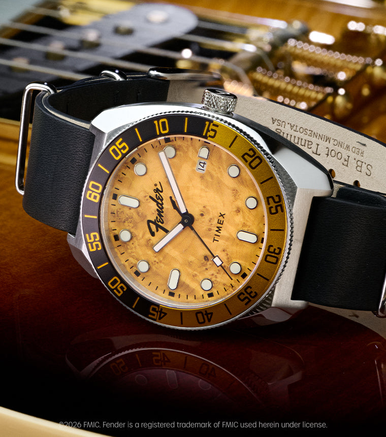 Fender x Timex Automatic watch with a silver-tone case, amber wood-grain dial, black-and-gold bezel, and black leather strap displayed on a Fender guitar.