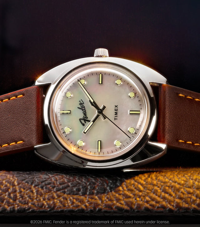 Fender x Timex watch with a silver-tone case, iridescent mother-of-pearl dial, and brown leather strap displayed on a guitar amplifier.