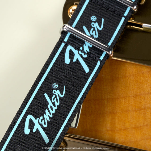 Close-up of a black Fender-branded strap with turquoise trim displayed beside an electric guitar.