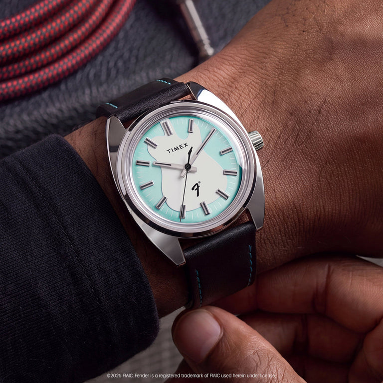 Fender x Timex watch worn on the wrist, featuring a silver-tone case, aqua dial accents, guitar motif, and black strap with turquoise stitching.