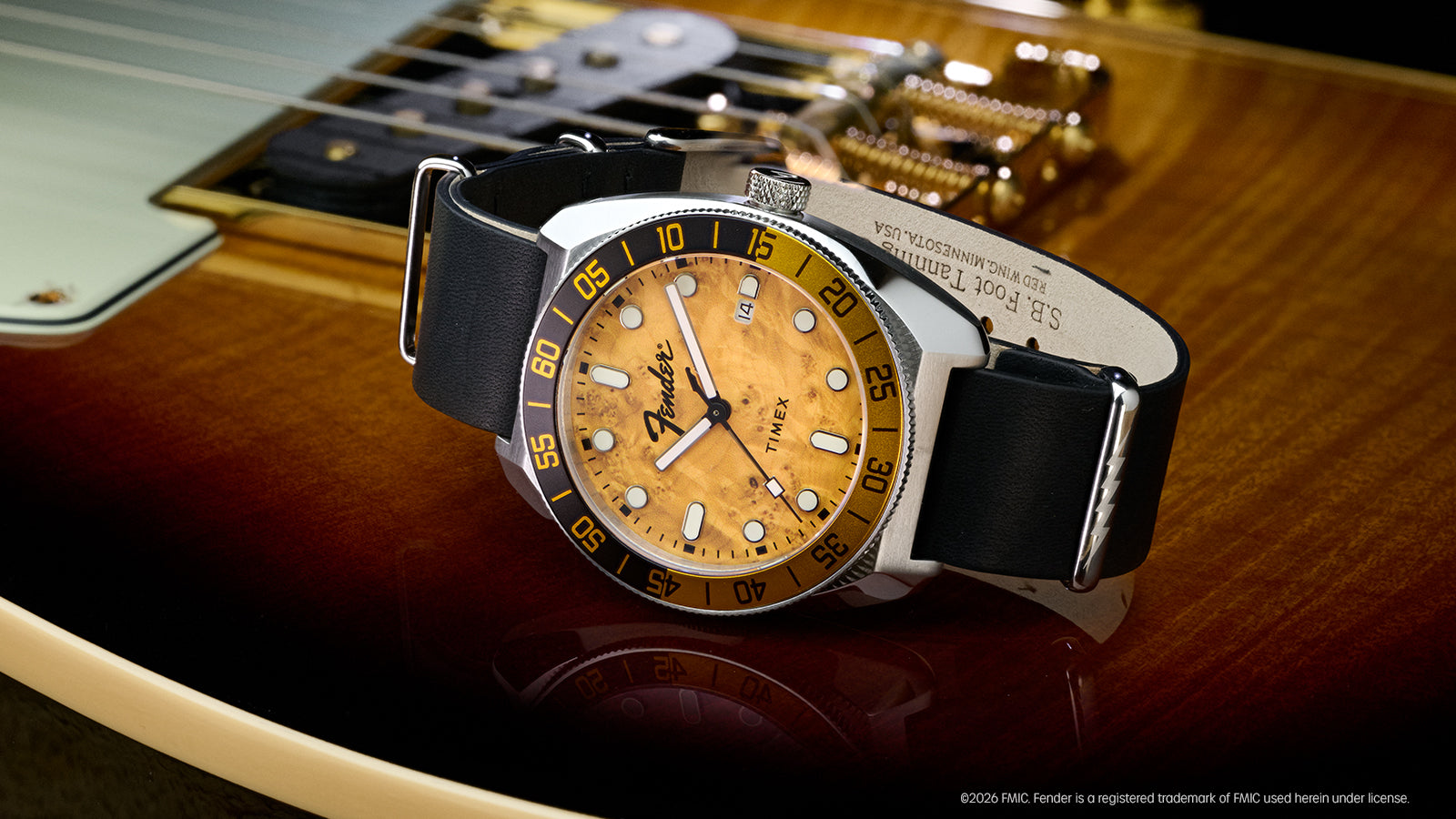 Fender x Timex Automatic watch displayed on a Fender electric guitar, featuring an amber wood-grain dial and black-and-gold bezel.